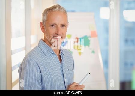 Uomo anziano in piedi accanto alla lavagna a fogli mobili nell'aula di formazione, tenendo in mano il poster arrotolato e indicando la mappa Foto Stock