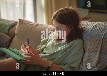 Concentrandosi sulla sua arte digitale, una ragazza preadolescente in abiti casual disegna su un tablet con uno stilo, godendosi il tempo libero mentre si rilassa in un intimo sof Foto Stock