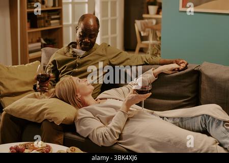 Coppia multietnica senior che si rilassa insieme sul divano e gusta il vino Foto Stock