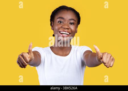 Donna africana gesturing Thumbs-Up con entrambe le mani su sfondo giallo Foto Stock