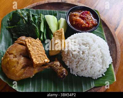 Tradizionale pasto indonesiano o nasi ayam lalapan composto da pollo fritto, tempeh, tofu, verdure, sambal e riso al vapore su un piatto di foglie di banana Foto Stock