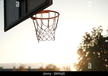 Un canestro da basket in un campo all'aperto catturato dalla calda luce del tramonto. La scena minimalista e nostalgica mette in risalto il cerchio e la rete contro Foto Stock