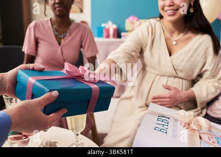 Giovane donna asiatica incinta sorridente mentre si avvicina alla scatola regalo dalle mani di un altro , seduta accanto alla giovane donna nera adulta durante la festa di rivelazione di genere Foto Stock