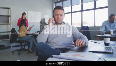 Uomo parlante che indossa un maglione grigio chiaro che fa telefonate alla scrivania dell'ufficio, con laptop e documenti Foto Stock