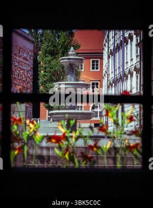 Un'affascinante fontana fissa un paesaggio urbano europeo, incorniciato dalle intricate finestre degli edifici circostanti e dalla vibrante sfocatura del fogliame in primo piano. Foto Stock