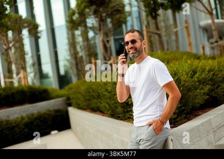 La luce del sole proietta un caldo bagliore mentre un uomo sorridente in un abbigliamento casual parla sul suo smartphone mentre si trova in un moderno spazio verde. Foto Stock
