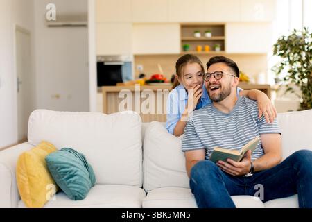 Un padre sorridente condivide una storia deliziosa con sua figlia, seduto su un comodo divano in una casa luminosa e moderna. Foto Stock