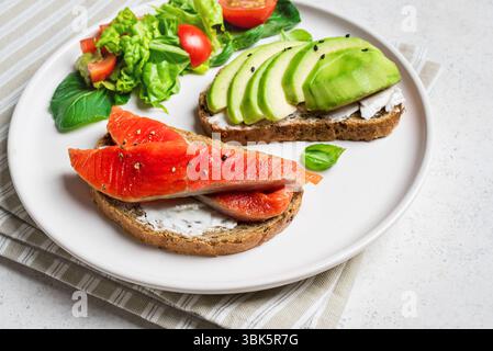 Pane tostato con avocado e salmone con formaggio spalmabile e insalata di verdure sul piatto da vicino. Colazione salutare, panini aperti con salmone e avocado. Foto Stock