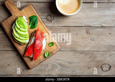 Toast all'avocado e al salmone con formaggio spalmabile e verdure, vista dall'alto. Colazione salutare, panini aperti con salmone e avocado e una tazza di caffè su Woode Foto Stock
