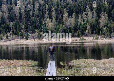 Paesaggio panoramico caratterizzato da una persona in piedi su un sentiero di legno vicino a un tranquillo lago circondato da pini Foto Stock