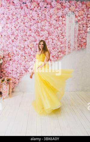 Una giovane donna caucasica con lunghi capelli marroni si intreccia in un abito giallo fluente. Si trova di fronte a una vivace parete di fiori rosa, creando un allegro Foto Stock