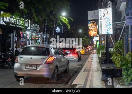 Bali, Indonesia - 25 dicembre 2024: Strada trafficata con molte auto e cartellone per donne. La strada è illuminata di notte Foto Stock