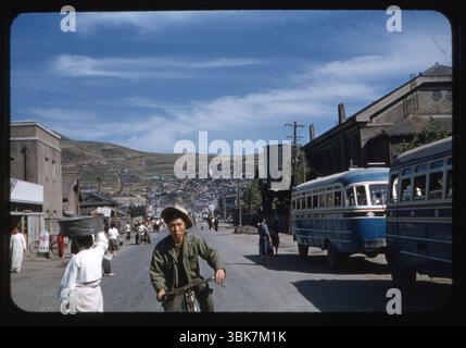 Vista sulla strada nel centro di Pusan (ora Busan), Corea del Sud, presa nell'ottobre 1956. Un uomo guida una bicicletta passando davanti a due autobus blu mentre i pedoni, alcuni in abito tradizionale coreano, camminano lungo la strada trafficata. Sullo sfondo sono visibili case in collina e pendii terrazzati. L'hotel si trova vicino all'ospedale svizzero, una struttura gestita da gruppi umanitari stranieri durante il periodo post-guerra di Corea. La scena cattura la trasformazione e la vita quotidiana della città poco dopo la fine della guerra. Questa fotografia è stata scattata dal Chief Petty Officer Wilfred K.C. Park, un coreano americano che serve nella Marina degli Stati Uniti. Foto Stock