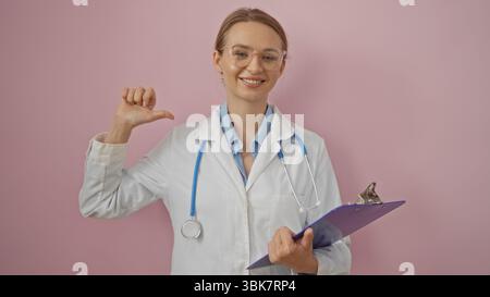 Medico donna che indica se stessa sorridendo con sicurezza con stetoscopio e appunti su sfondo rosa isolato Foto Stock
