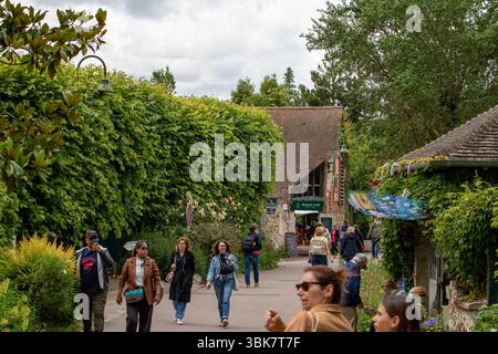 Una scena di strada con turisti che camminano davanti a un caffè e una vegetazione lussureggiante sotto un cielo nuvoloso a Giverny, in Francia. Foto Stock