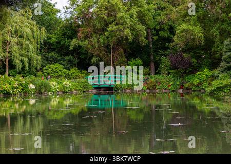 Il verde rigoglioso incornicia un laghetto riflettente con una passerella verde ad arco sullo sfondo a Giverny, Francia. Foto Stock