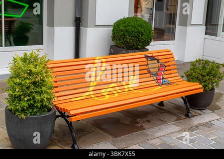 Una panchina arancione vibrante con la parola "amore" e un'illustrazione a cartoni animati si trova su un sentiero in mattoni, fiancheggiato da piante verdi in vaso al la Vallee Village, Fran Foto Stock