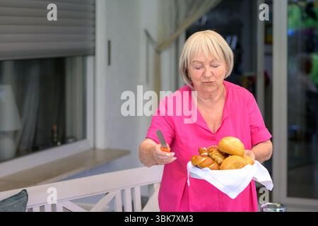 Una donna anziana con una camicetta rosa porta un cesto pieno di panini e pretzel freschi, tenendo un coltello per il pane nell'altra mano. Foto Stock