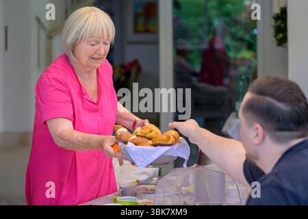 Una donna anziana con una camicetta rosa brillante offre un cestino di pretzel tradizionali a un uomo seduto durante una riunione di famiglia all'aperto. Foto Stock