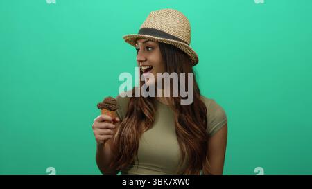 Donna che si gusta un gelato su uno sfondo verde, indossa un cappello e sorride, ritraendo un momento vivace con un dessert al cioccolato. Foto Stock