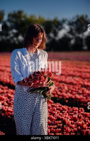 Elegante donna in polka dots si erge tranquillamente in un campo di tulipani, tenendo freschi fiori rossi e crogiolandosi nella luce dorata dell'ora. Foto Stock