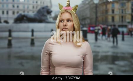 Una donna con diadema unicorno si trova in una strada cittadina che sembra divertente e preoccupata, sullo sfondo della vita urbana, trasmettendo un contrasto stravagante tra Foto Stock