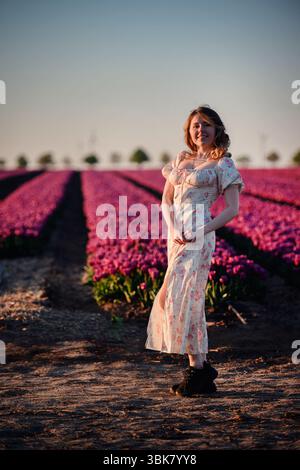 Le donne si godono il tramonto in un campo di tulipani in fiore, indossando un abito floreale e scarpe nere, inondate da una calda luce dorata. Foto Stock