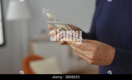 Uomo maturo che conta dollari americani in una casa accogliente, evidenziando pianificazione finanziaria, risparmi e budget in una finanza personale interna Foto Stock