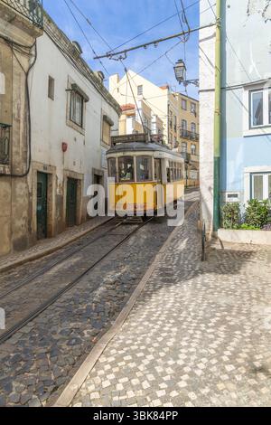 Tram giallo d'epoca su una strada acciottolata di Lisbona, che si snoda attraverso colorati edifici storici sotto un cielo luminoso. Un classico simbolo della città. Foto Stock