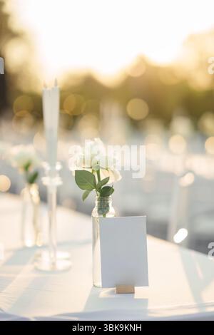 Elegante tavolo per matrimoni all'aperto con cartello bianco, fiore di rosa bianco e sedie bianche in giardino durante le feste nuziali Foto Stock