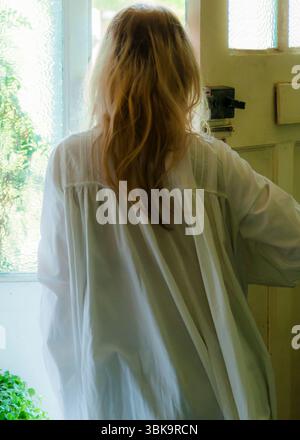 Una donna con lunghi capelli biondi sta in una porta, guardando fuori da una finestra. Sta indossando un abito da notte bianco. Foto Stock