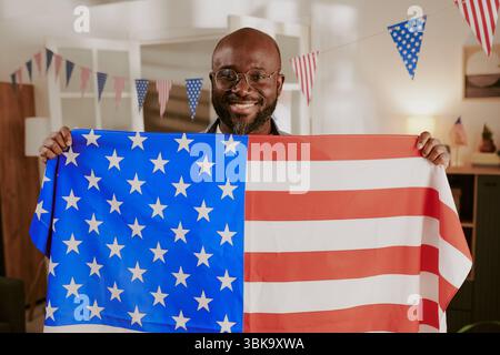Ritratto dell'uomo Nero di mezza età sorridente mentre tiene la bandiera americana in casa Foto Stock