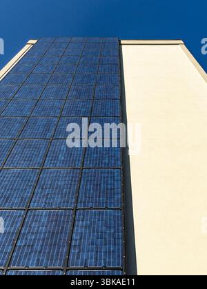 Vista dal basso dei pannelli solari verticali sulla facciata di un moderno edificio urbano sotto il cielo blu, che illustra l'energia pulita e lo sviluppo sostenibile della città. Foto Stock