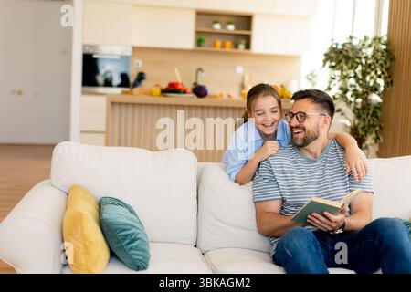 Un padre sorridente condivide una storia deliziosa con sua figlia, seduto su un comodo divano in una casa luminosa e moderna. Foto Stock