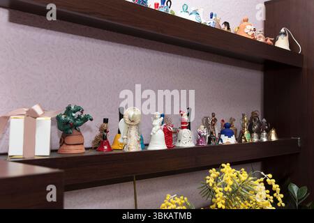 Collezione di campane decorative souvenir in ceramica e metallo su scaffali in legno scuro, fotografate all'interno su pareti in lilla testurizzata Foto Stock