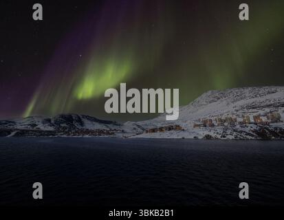 Aurora boreale sulla città costiera di Nuuk, in Groenlandia, con edifici innevati e montagne in inverno. Gli archi di Aurora attraversano il cielo notturno artico. Foto Stock