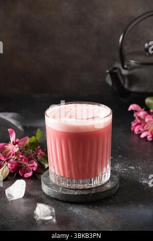 Latte matcha rosa con fiori sakura decorati a polvere su sfondo scuro. Bevanda alla moda. Rinfrescante matcha rosa ghiacciato. Verticale Foto Stock