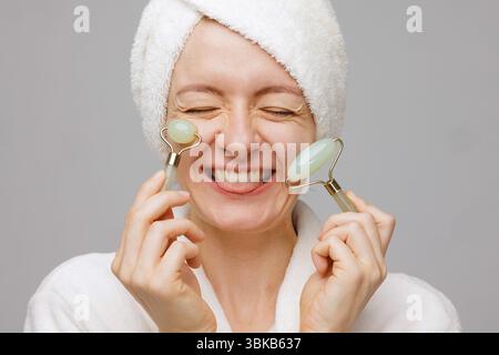 Donna felice in asciugamano e accappatoio che utilizza rulli facciali in giada per la cura della pelle, levigatura delle rughe e ringiovanimento nella spa di casa. Foto Stock