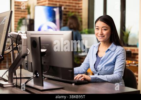 Donna creativa asiatica seduta alla scrivania dell'ufficio, usando la tastiera del computer e sorridendo alla fotocamera. Ritratto di una donna d'affari che lavora su un pc desktop, digita, risponde alle e-mail aziendali nello spazio di lavoro a muro. Foto Stock