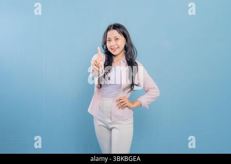 Una donna è in piedi di fronte a un muro blu con una camicia rosa e pantaloni bianchi. Sta puntando il dito verso l'alto e sorridendo Foto Stock