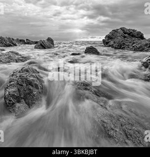 Un primo piano grandangolare di Un'onda che scorre a riva sulle Sea Rocks in Una spettacolare immagine verticale a contrasto elevato in bianco e nero Foto Stock