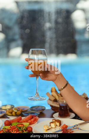 Un tavolo in legno preparato per la colazione in hotel con insalata, olive, pomodori e formaggio, mentre la mano di una donna regge un bicchiere di succo di frutta. Una piscina tranquilla e' vi Foto Stock