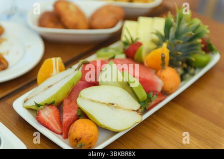 Un vivace mix di frutta fresca, tra cui ananas, fragole, anguria, pera e cachi, esposto su un ampio piatto in cima a un rustico tavolo in legno i. Foto Stock