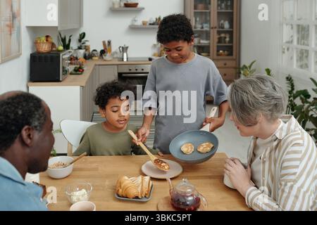 La famiglia condivide la colazione con pancake in un'accogliente cucina. Vecchio tavolo in legno, moderni accessori da cucina, luce naturale che crea un'atmosfera calda. Ragazzo che serve pancake Foto Stock