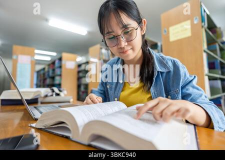 Una giovane donna asiatica studiosa che indossa gli occhiali si siede a una scrivania con un laptop e legge un grande libro, concentrandosi sulla sua ricerca per un assegnatario scolastico Foto Stock