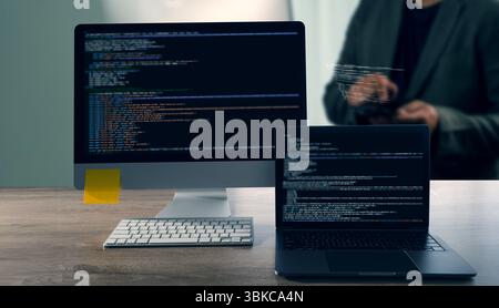Un codice di programmazione che rappresenta il primo piano di un monitor per computer e di un laptop che visualizza il codice, con sfondo della persona che utilizza un tablet, un moderno spazio di lavoro Foto Stock