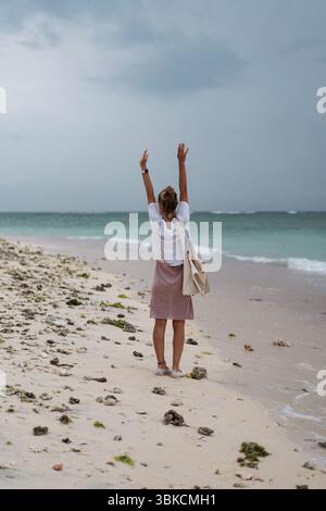 Giovane donna in piedi con le braccia sollevate sulla spiaggia tropicale rocciosa Foto Stock