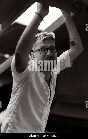 Uomo sicuro di sé in camicia di lino che posa sotto travi di legno Foto Stock