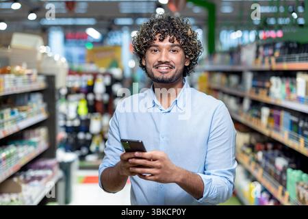 Un uomo sorridente in un supermercato, che tiene un telefono, guarda la macchina fotografica. Gli scaffali con prodotti sono sullo sfondo. Foto Stock