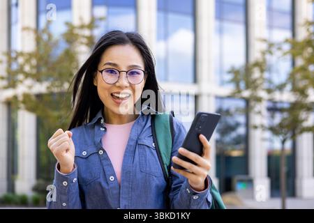 Una donna asiatica eccitata che indossa gli occhiali tiene un telefono e festeggia con il pugno sollevato fuori, vicino a un edificio. Foto Stock